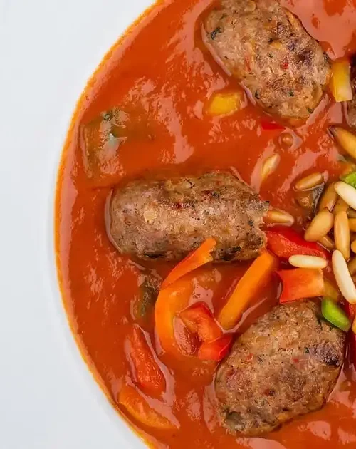 Involtini di agnello al sugo su piatto bianco, guarniti con pinoli e prezzemolo