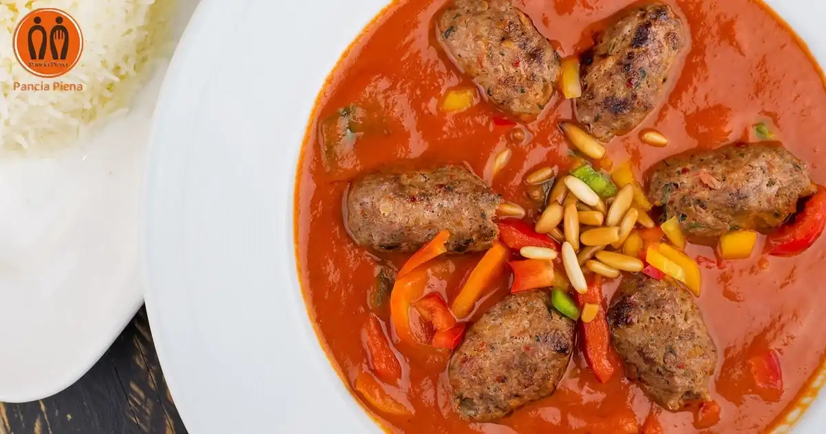 Involtini di agnello al sugo su piatto bianco, guarniti con pinoli e prezzemolo